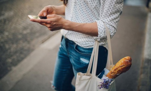 woman walks down the street and texts with a bag of groceries on her arm